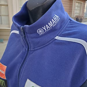 May include: A blue Yamaha Factory Racing zip-up jacket with white accents. The jacket features the Yamaha logo on the collar and various sponsor patches, including Monster Energy and Movistar. The jacket is displayed on a black mannequin.