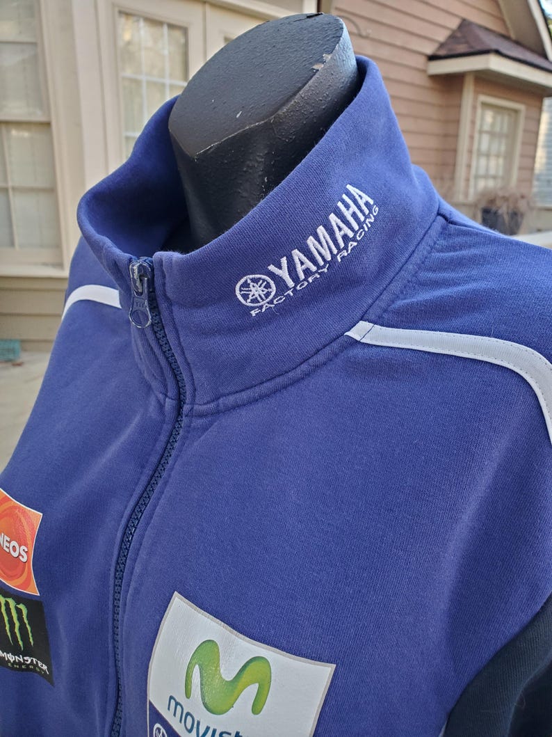 May include: A blue Yamaha Factory Racing zip-up jacket with white accents. The jacket features the Yamaha logo on the collar and various sponsor patches, including Monster Energy and Movistar. The jacket is displayed on a black mannequin.