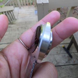 May include: A silver-toned wristwatch with a black face and a rectangular, brushed metal case. The watch has a matching silver band with a clasp. The watch is held in a hand, with a ring on the finger.