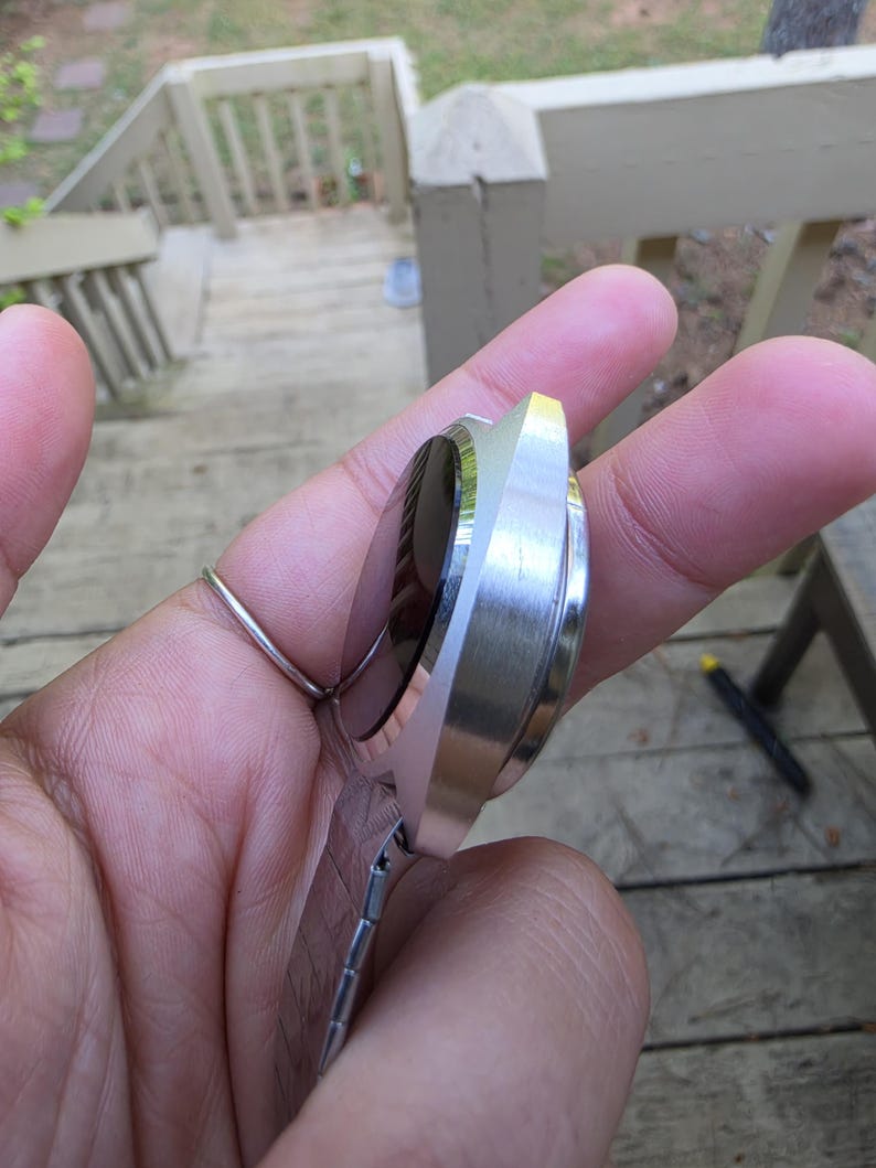 May include: A silver-toned wristwatch with a black face and a rectangular, brushed metal case. The watch has a matching silver band with a clasp. The watch is held in a hand, with a ring on the finger.