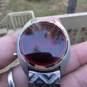May include: A vintage silver-toned digital watch with a red display. The watch face is round with a silver bezel and a red border. The watch band is silver and has a geometric pattern. The watch is being held up in front of a blurred outdoor background.