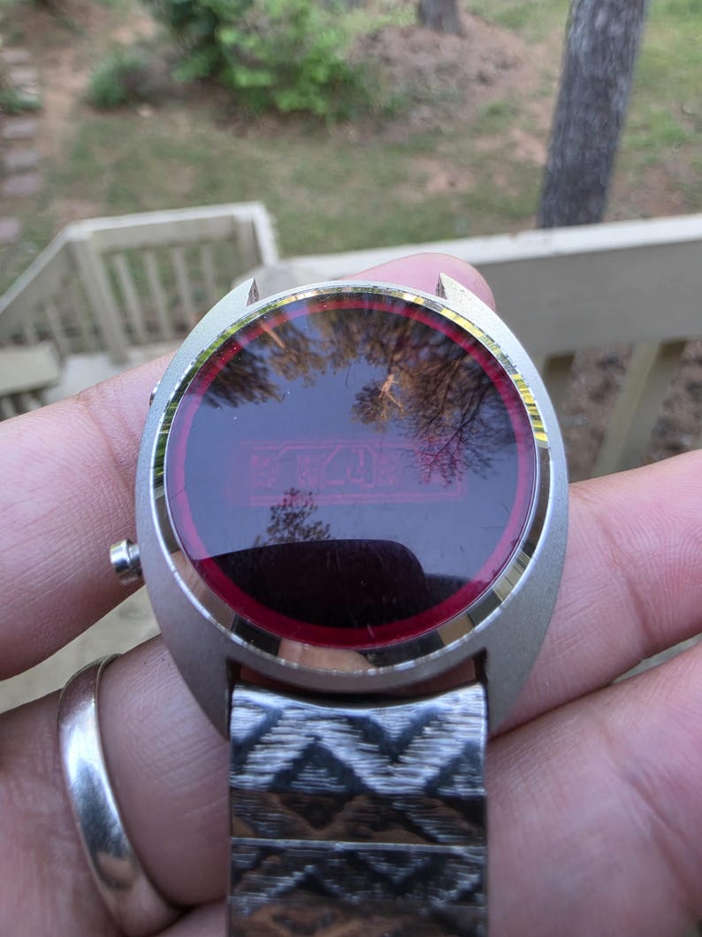 May include: A vintage silver-toned digital watch with a red display. The watch face is round with a silver bezel and a red border. The watch band is silver and has a geometric pattern. The watch is being held up in front of a blurred outdoor background.
