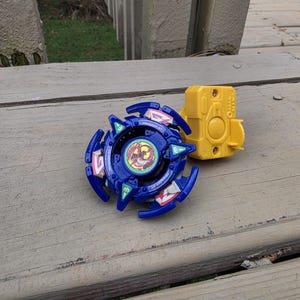 May include: A blue spinning top toy with a central circular emblem featuring a red and yellow graphic, surrounded by white and green accents. Next to the spinning top is a yellow plastic handheld launcher with a central circular button.
