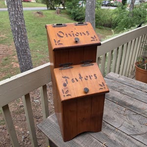 May include: A wooden storage bin with two compartments, labeled "Onions" and "Taters". The bin is a warm brown color with decorative leaf cutouts and small knobs. The bin is sitting on a wooden surface.