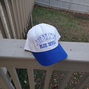May include: A white baseball cap with a blue brim and the word "DUKE" embroidered in blue. The text "BLUE DEVILS" is also embroidered on the cap. The cap is resting on a white railing.