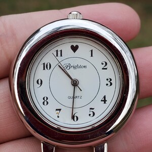 May include: A silver-toned Brighton quartz watch face with a white dial. Black numerals mark the hours, with a heart symbol above the 11. The word "Brighton" is printed below the heart, and "QUARTZ" is printed below the center.