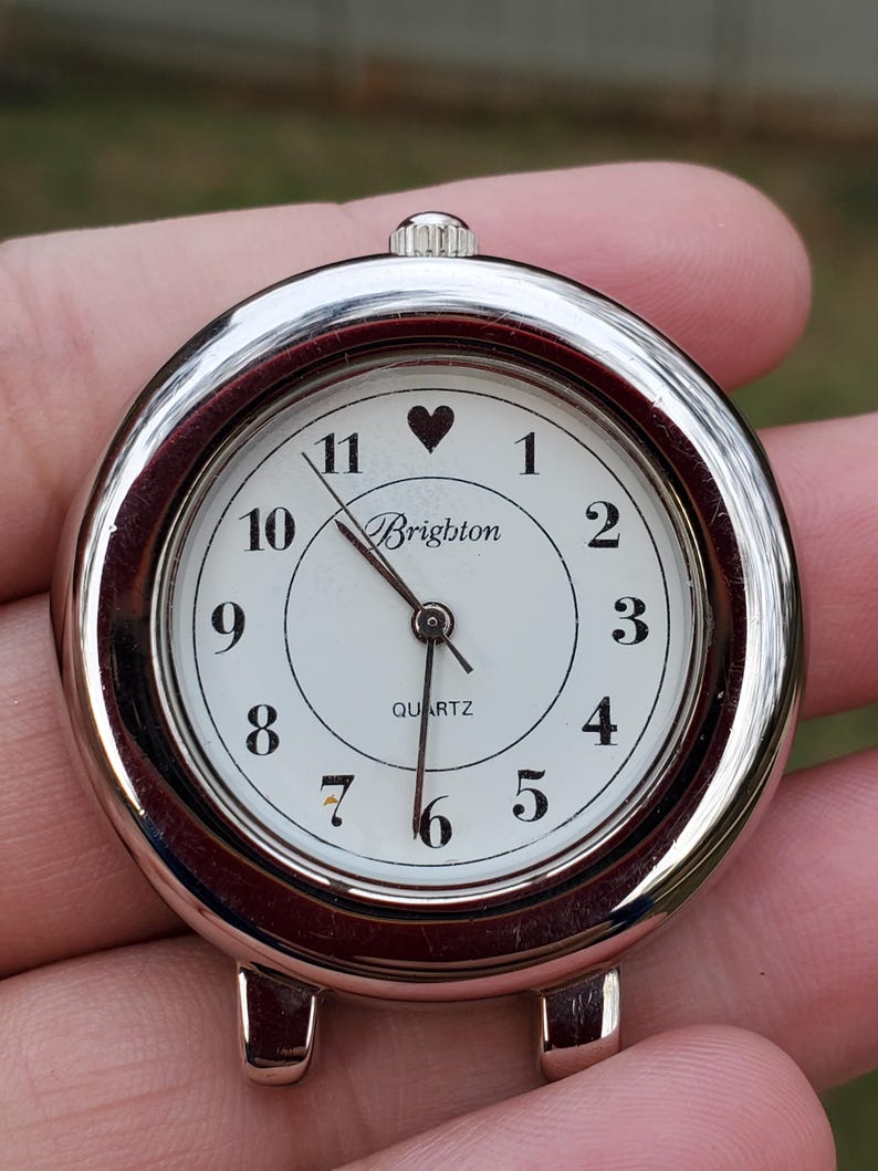 May include: A silver-toned Brighton quartz watch face with a white dial. Black numerals mark the hours, with a heart symbol above the 11. The word "Brighton" is printed below the heart, and "QUARTZ" is printed below the center.