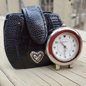 May include: A black leather watch case with a silver heart-shaped accent and a matching black strap. A silver-toned Brighton quartz watch with a white face, red bezel, and a heart at the 12 o'clock position is next to the case.
