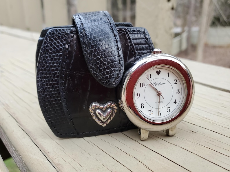 May include: A black leather watch case with a silver heart-shaped accent and a matching black strap. A silver-toned Brighton quartz watch with a white face, red bezel, and a heart at the 12 o'clock position is next to the case.