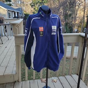 May include: A blue zip-up jacket with white and black accents. The jacket features sponsor logos and the Movistar logo on the collar. The jacket is displayed on a black mannequin stand on a wooden deck.