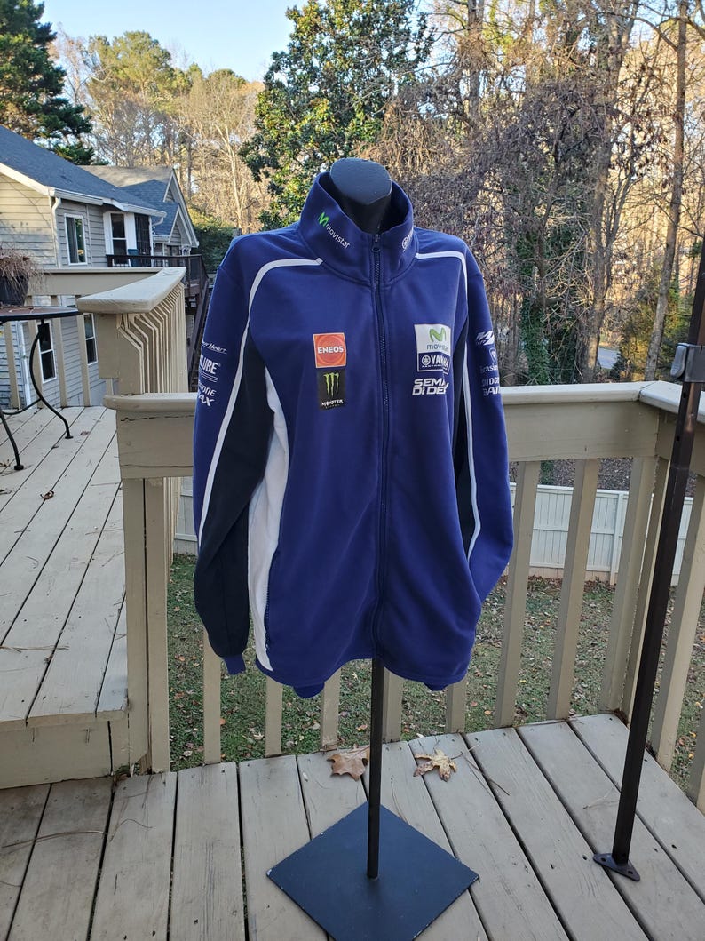 May include: A blue zip-up jacket with white and black accents. The jacket features sponsor logos and the Movistar logo on the collar. The jacket is displayed on a black mannequin stand on a wooden deck.