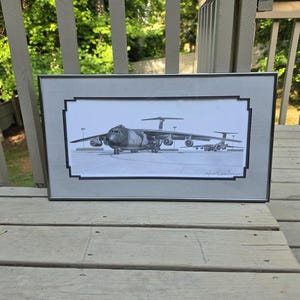 May include: A framed black and white pencil drawing of a large military cargo plane on a runway. The artwork is in a silver frame with a decorative border. The plane has multiple engines and a high tail.