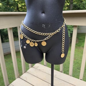 May include: A gold-toned chain belt featuring two layered strands of oval links. The lower strand is adorned with five circular coin-shaped charms of varying sizes that hang from the chain, and one end of the belt hangs down as a long chain.
