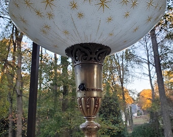 May include: A vintage floor lamp with a white, star-patterned shade. The shade has gold starburst designs. The lamp features a brass-colored base and stem with ornate detailing. The lamp is outdoors, with trees in the background.