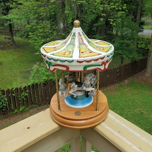 May include: A porcelain carousel with three horses, a clown, and a blue and white base. The carousel is painted with yellow, green, red, and white details. The carousel is on a wooden base.