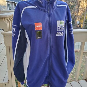 May include: A blue Yamaha zip-up jacket with white and black accents. The jacket features multiple sponsor logos, including "Movistar," "Eneos," and "Monster." The jacket has a high collar and a full-length zipper.
