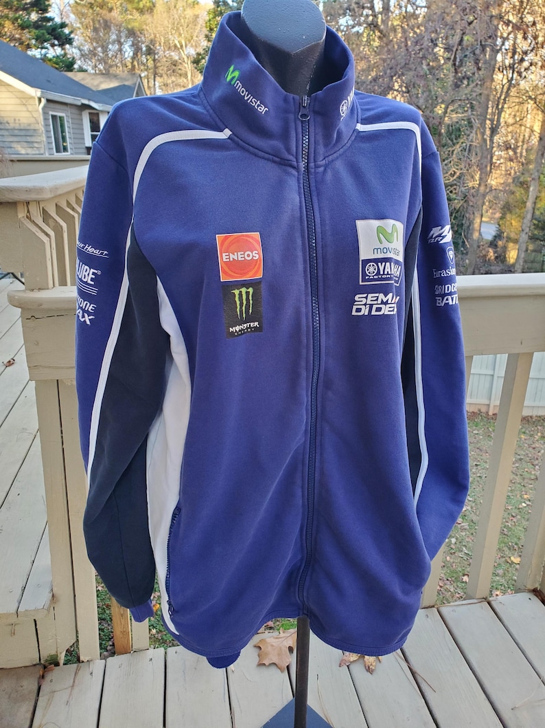 May include: A blue Yamaha zip-up jacket with white and black accents. The jacket features multiple sponsor logos, including "Movistar," "Eneos," and "Monster." The jacket has a high collar and a full-length zipper.