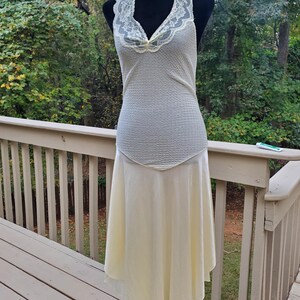 Puede incluir: Un vestido sin mangas de color marfil con un escote de encaje y una falda fluida. El vestido est&aacute; en un maniqu&iacute; y se muestra contra una plataforma de madera.