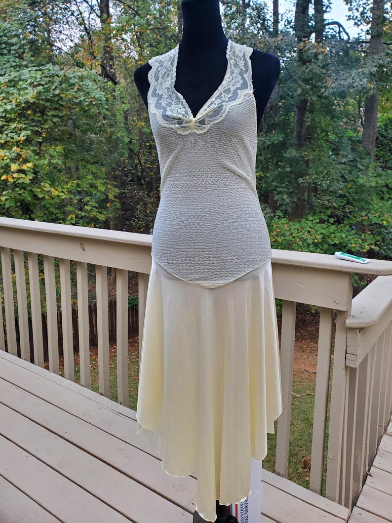 Puede incluir: Un vestido sin mangas de color marfil con un escote de encaje y una falda fluida. El vestido est&aacute; en un maniqu&iacute; y se muestra contra una plataforma de madera.