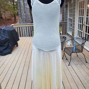 Puede incluir: Un vestido blanco sin mangas con una superposici&oacute;n de encaje en los hombros y una falda fluida. El vestido est&aacute; en un maniqu&iacute; y se fotograf&iacute;a contra una plataforma de madera.