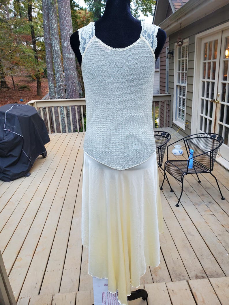 Puede incluir: Un vestido blanco sin mangas con una superposici&oacute;n de encaje en los hombros y una falda fluida. El vestido est&aacute; en un maniqu&iacute; y se fotograf&iacute;a contra una plataforma de madera.