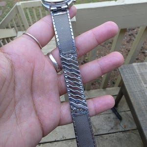 May include: A vintage silver-toned wristwatch with a mesh band. The watch face is round with a light-colored dial. The band features a decorative, patterned section with the words "Stainless Steel".