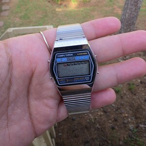 May include: A silver-toned digital watch with a black face and a metal band. The watch face displays the text "COMPU CHRON" and the days of the week. The watch is held in a hand.