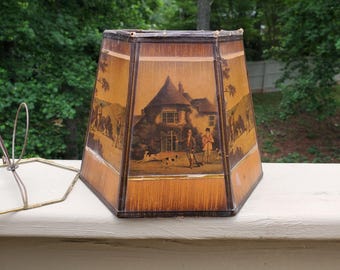 May include: A vintage lampshade with a hexagonal shape. The shade features a detailed illustration of a house and figures in a landscape, framed by a brown and gold border. The background shows a green outdoor setting.