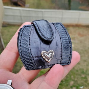 May include: Black, rectangular, small wallet with a textured, reptile-like pattern. The wallet has a silver heart-shaped accent and a clasp closure. The wallet is held in a person's hand.