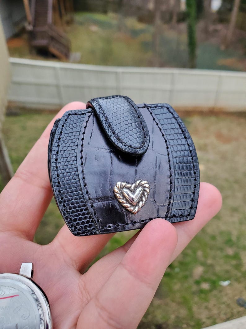 May include: Black, rectangular, small wallet with a textured, reptile-like pattern. The wallet has a silver heart-shaped accent and a clasp closure. The wallet is held in a person's hand.