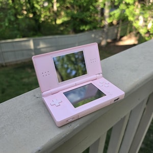 May include: A light pink Nintendo DS handheld game console, open and resting on a wooden railing. The console features two screens, a directional pad, and action buttons. The background shows a blurred view of trees and a fence.
