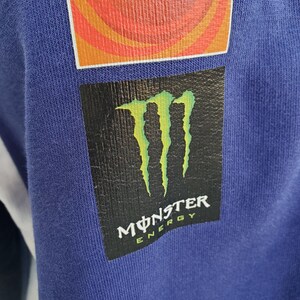 May include: Close-up of a blue fabric with two patches. The top patch is orange and red with the word "ENEOS" in white. The bottom patch is black with a green claw design and the words "MONSTER ENERGY" in white.