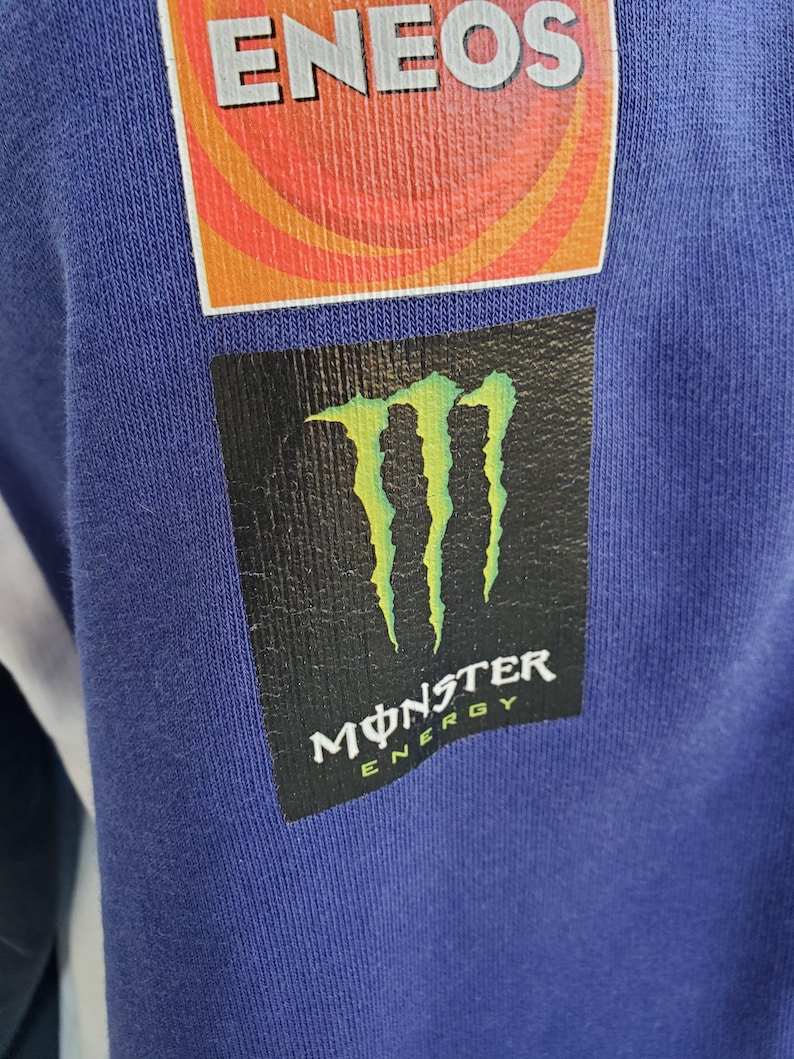 May include: Close-up of a blue fabric with two patches. The top patch is orange and red with the word "ENEOS" in white. The bottom patch is black with a green claw design and the words "MONSTER ENERGY" in white.