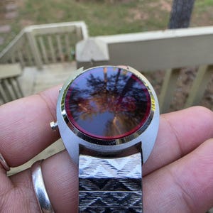May include: A vintage silver-toned wristwatch with a round face and a red-tinted display. The watch has a patterned metal band. The watch is held in a hand, with a silver ring on a finger.