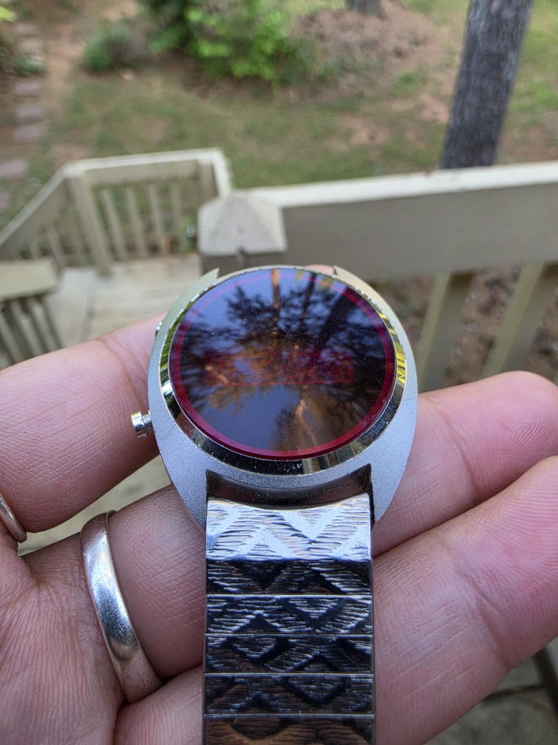 May include: A vintage silver-toned wristwatch with a round face and a red-tinted display. The watch has a patterned metal band. The watch is held in a hand, with a silver ring on a finger.