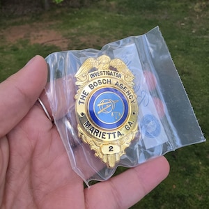 May include: A gold-coloured investigator badge from The Bosch Agency, Marietta, GA, with a blue and white centre. The badge is in a clear plastic bag. The badge has the number 2.