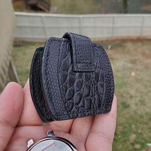 May include: A black watch travel case with a textured, reptile-like pattern. The case is closed with a flap and buckle. A silver watch face is visible below the case, with the words "TIME TOTES" printed on the watch face.