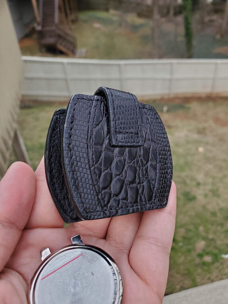 May include: A black watch travel case with a textured, reptile-like pattern. The case is closed with a flap and buckle. A silver watch face is visible below the case, with the words "TIME TOTES" printed on the watch face.