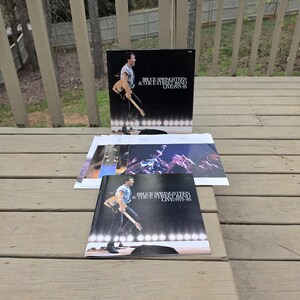 Puede incluir: Una portada de álbum de vinilo con Bruce Springsteen & The E Street Band Live 1975-85. La portada muestra a Springsteen tocando la guitarra en un escenario. El álbum se muestra en una terraza de madera con otros insertos.