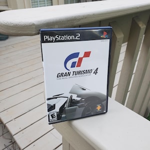 Peut inclure: Une boîte de jeu PlayStation 2 pour Gran Turismo 4, le simulateur de conduite réaliste. La boîte est noire avec un logo blanc et rouge et une image d'une voiture de sport argentée avec sa porte ouverte.