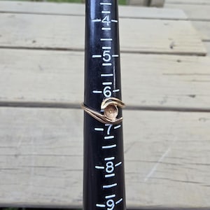 May include: A gold-toned metal ring with a bypass-style band and a central circular setting for a stone, positioned on a black plastic ring mandrel marked with white size numbers, showing the ring rests at approximately a size 6.5 on the scale.