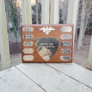 May include: A wooden plaque with a gold shield in the center that reads "HOSTESS CAKE Salesman of the Year". The plaque has several smaller gold plaques with names and dates on them.