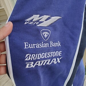 May include: Close-up of a blue athletic garment with white and black accents. The garment features embroidered logos and text, including "VZR", "Eurasian Bank", "Bridgestone", and "Battlax". The fabric appears to be a soft, knit material.
