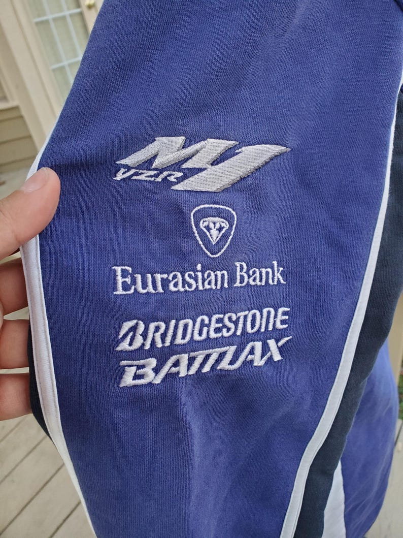 May include: Close-up of a blue athletic garment with white and black accents. The garment features embroidered logos and text, including "VZR", "Eurasian Bank", "Bridgestone", and "Battlax". The fabric appears to be a soft, knit material.