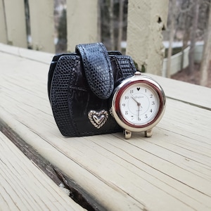 May include: A black leatherette travel clock case with a silver-tone Brighton clock. The clock has a white face with black numerals and hands. A silver heart is on the case. The case is closed with a flap.