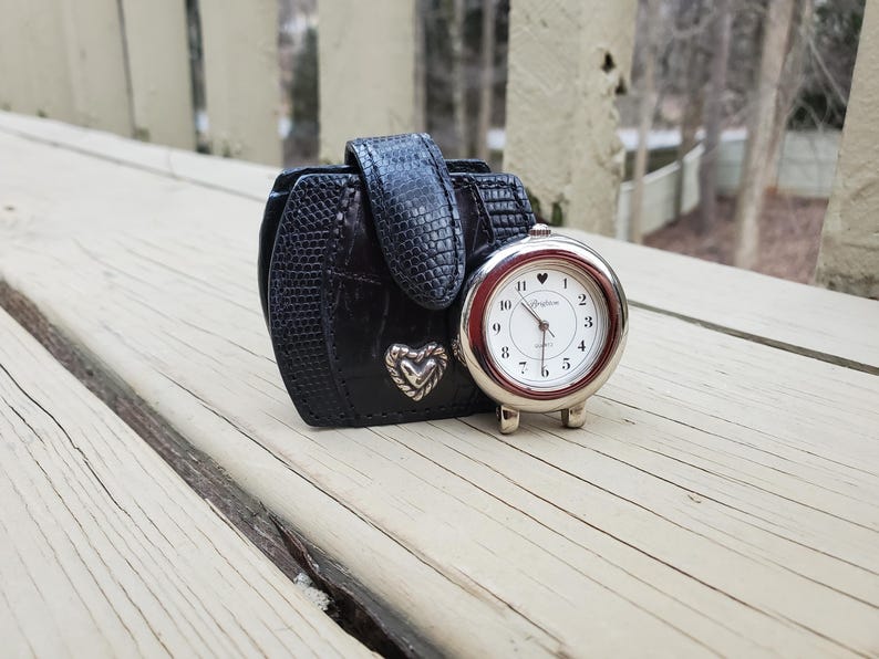May include: A black leatherette travel clock case with a silver-tone Brighton clock. The clock has a white face with black numerals and hands. A silver heart is on the case. The case is closed with a flap.
