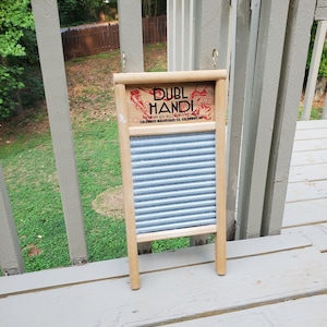 Op de afbeelding: Een houten wasbord met een gegolfd metalen oppervlak. Het wasbord heeft een rood en wit label met de tekst "DUBL HANDI" en "WASH EITHER SIDE ACCORDING TO WEAR". Het label bevat ook de tekst "COLUMBUS WASHBOARD CO. COLUMBUS, OH."