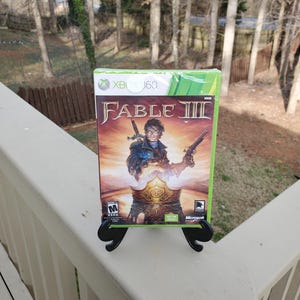 May include: A video game case for Fable III for the Xbox 360. The case is green and black with a picture of a man holding a sword and a gun. The game is rated M for Mature.