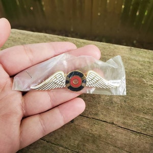 May include: A gold-colored pin with white wings and a red, black, and white circular design in the center.