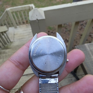 May include: A stainless steel watch with a silver mesh band. The watch face is silver with the text "BASE METAL STAINLESS STEEL BACK U.S.A." and the number "451323". The watch is held in a hand.